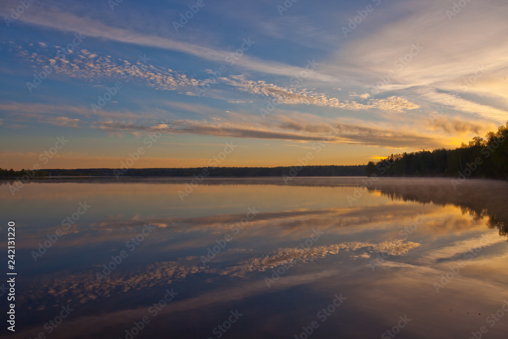 Dawn over the quiet water surface of the lake. The morning blue sky is lit by the orange light of the sun.