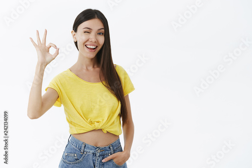 Good-looking female fitness trainer assuring she will help winking joyfully showing okay or excellent gesture with raised fingers holding hand in pockets standing confident, delighted over grey wall