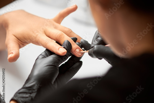 Close-up shot of beautician hands in black rubber gloves applying with brush transparent gel polish on woman fingernails on blurred background. Healthcare  beauty cosmetics  spa procedure concept.