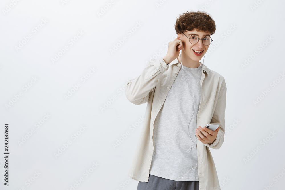 Indoor shot of charismatic happy and carefree young european male model with curly hairstyle in round glasses putting on earbud as listening music with app in smartphone smiling at camera satisfied Indoor shot of charismatic happy and carefree young european male model with curly hairstyle in round glasses putting on earbud as listening music with app in smartphone smiling at camera satisfied
