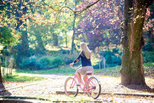 Side view of young active blond long-haired woman in skirt and blouse cycling pink lady bicycle along park paved alley with fallen golden leaves on blurred colorful foggy bokeh foliage background.