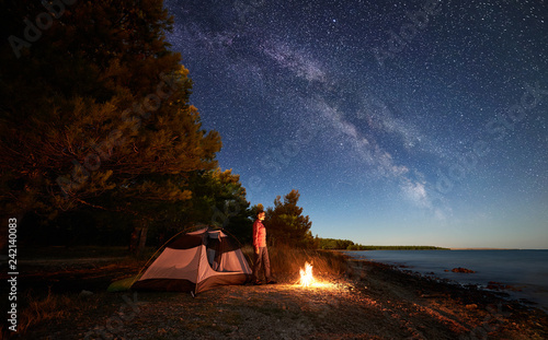 Night camping at sea coast. Female hker standing at campfire near tourist tent and forest, enjoying beautiful view of starry sky with Milky way and clear blue water. Tourism, active lifestyle concept