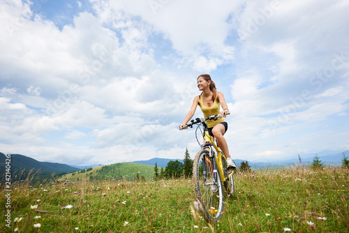 Attractive happy female cyclist riding on yellow mountain bike on a grassy hill, enjoying summer day in the mountains. Outdoor sport activity, lifestyle concept