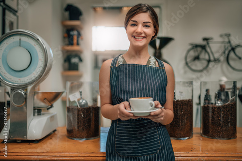 Young beautiful waitress smiling serving coffee with latte art while standing in coffee shop.