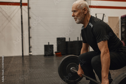 Side view of confident senior male bodybuilder with gray beard doing squats indoors holding barbell. Attractive muscular retired man exercising at fitness center, shaping up, gaining srong body