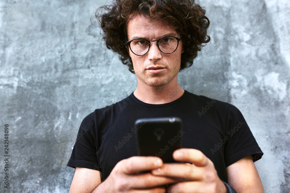 Image of handsome smart man standing outdoors texting on mobile phone. Young male with curly hair wears spectacles resting outside in the city browsing on his cell phone on concrete gray background. Image of handsome smart man standing outdoors texting on mobile phone. Young male with curly hair wears spectacles resting outside in the city browsing on his cell phone on concrete gray background.