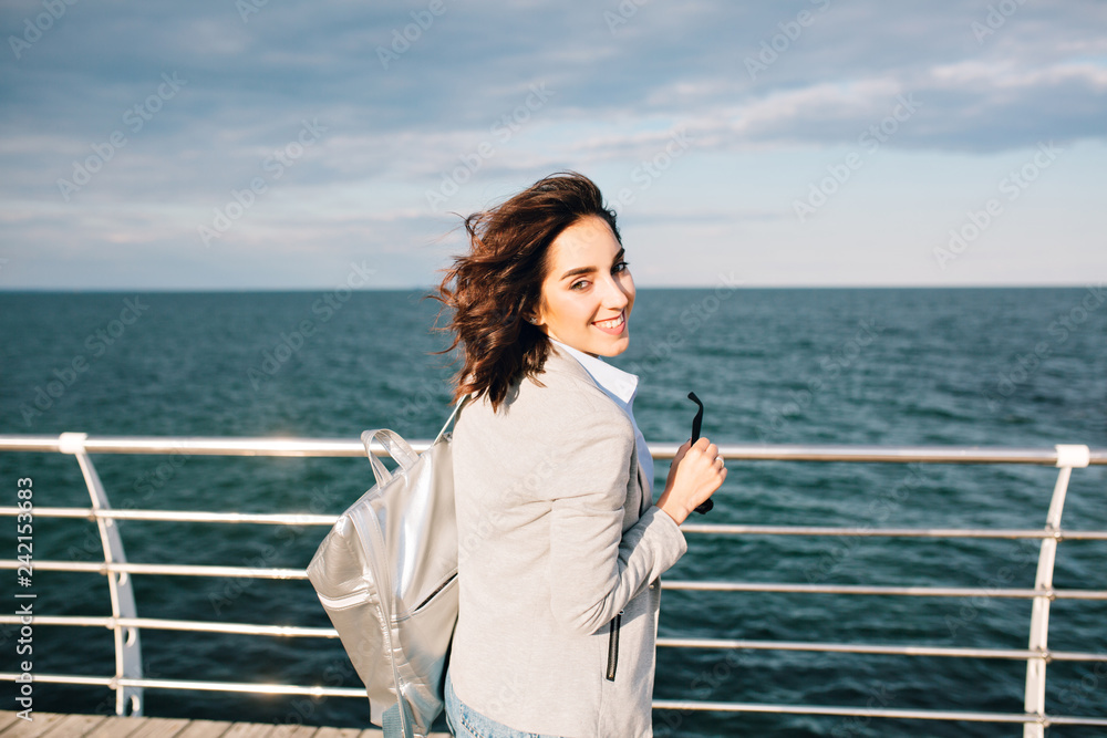 Portrait of cute brunette girl with short hair smiling to the camera on sea background. She wears shirt, jacket and bag. View from back. Portrait of cute brunette girl with short hair smiling to the camera on sea background. She wears shirt, jacket and bag. View from back.