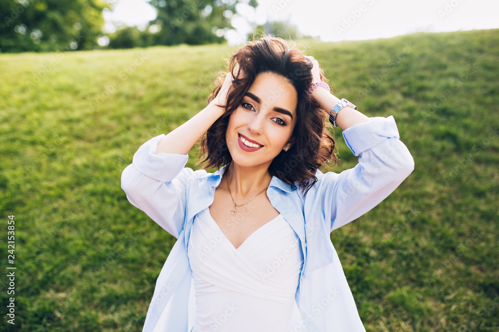 Portrait of cute brunette girl with short hair posing to camera in park. She wears white T-shirt and blue shirt. Portrait of cute brunette girl with short hair posing to camera in park. She wears white T-shirt and blue shirt.