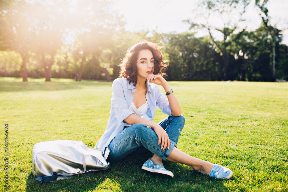 Full-length photo of cute brunette girl with short hair sitting on grass in sunlight in park . She wears white T-shirt, shirt and jeans, shoes, bag. Full-length photo of cute brunette girl with short hair sitting on grass in sunlight in park . She wears white T-shirt, shirt and jeans, shoes, bag.