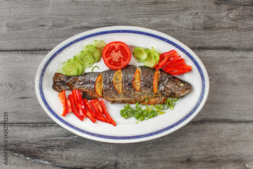 Tabletop view, grilled trout on oval plate, orange slices, tomatoes, pepper, cucumber and green onion around.