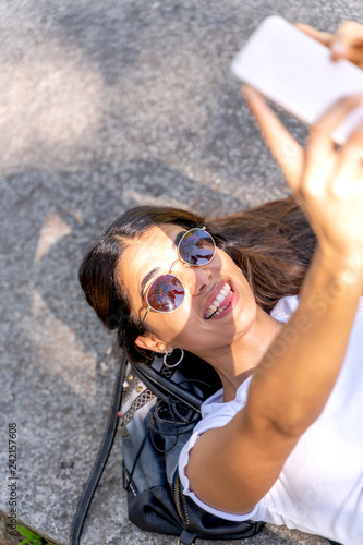 A beautiful young woman in a white shirt and sunglasses taking selfie and making funny faces while lying on a concrete surface.