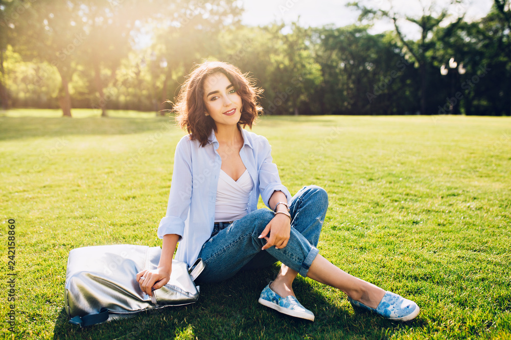 Cute brunette girl with short hair is sitting on grass in sunlight in park . She wears white T-shirt, shirt and jeans, shoes, bag. She looks happy to the camera. Cute brunette girl with short hair is sitting on grass in sunlight in park . She wears white T-shirt, shirt and jeans, shoes, bag. She looks happy to the camera.