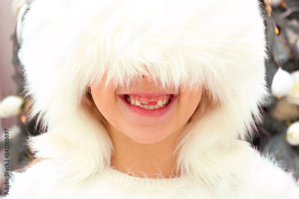 Cropped Shot Of A Smiling Girl Wearing A White Hat Over Christmas Tree Background. Face Of Little Girl. Happiness, Childhood, Christmas, Winter, Holidays Concept. Happy Toothless Girl Portrait.
