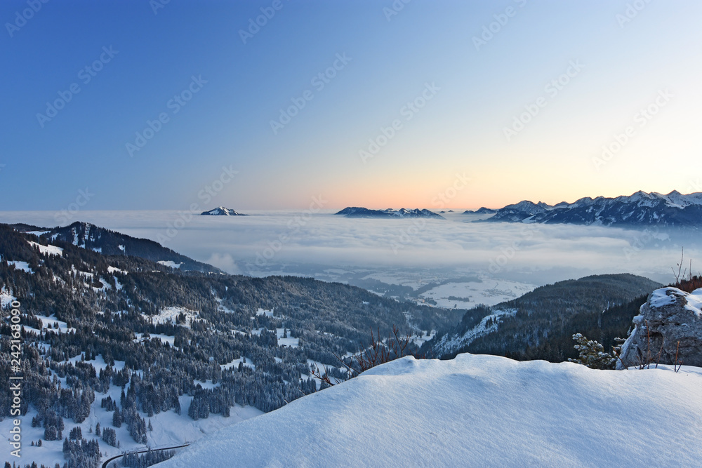 Beautiful morning mood in the Allgaeu Alps at a cold winter day. Snow-covered alpine landscape with mountains sticking out of a cloud layer and red glowing horizon before sunrise (Bavaria, Germany).