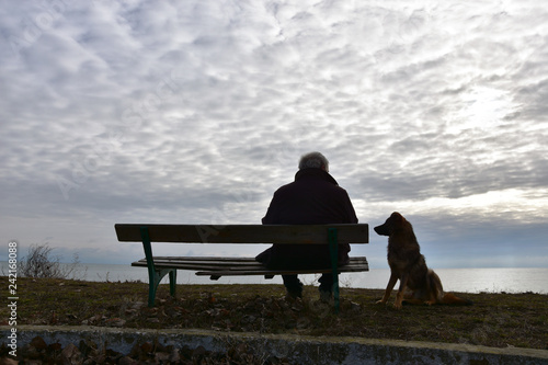 Elderly man sitting on an old wooden bench above the see in a cloudy winter day, his dog quietly standing by. Nostalgic mood. Time is passing by