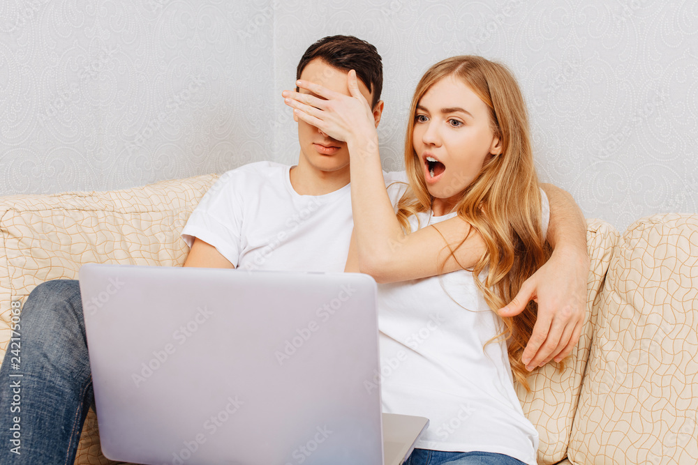 Beautiful young couple, man and woman, using laptop, shocked girl closes eyes to man, sitting at home on sofa