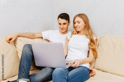 Beautiful young couple, man and woman using a laptop, hugging and smiling, sitting at home on the couch