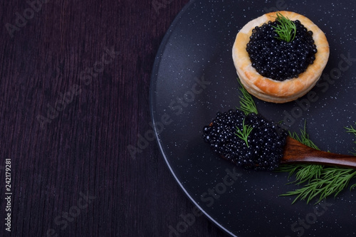 Black caviar in a wooden spoon and puff pastry tartlet on a dark plate