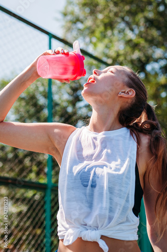 Portrait of sporty young sexy woman drinking cool water from  bottle on a summer sports field outdoors..