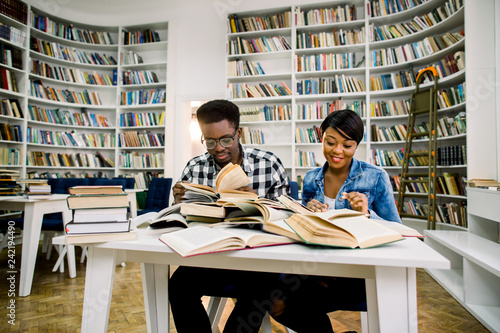 Cheerful african american students boy and girl sitting in university campus with books and learning for exams, multiracial hipster girl and boy smiling and talking while reading books in library