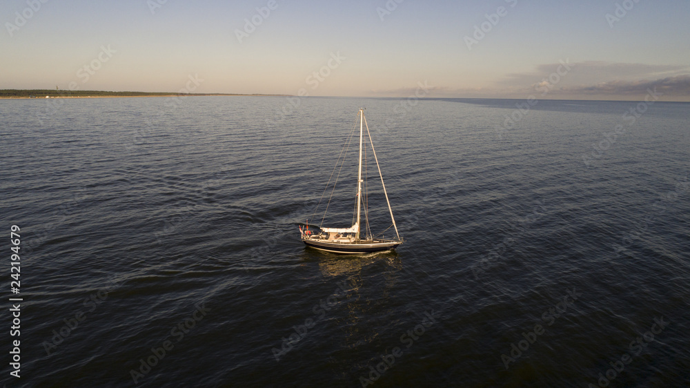 Fototapeta premium Aerial shot from the middle of the sea of lonely sailboat with coiled sails floats on a calm sea. Sunrise in summer or spring. In the background sandy coast, beach hotel and canal.