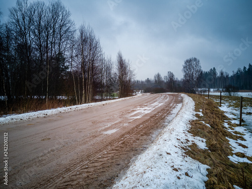 Empty Countryside Landscape in Cloudy Winter Day with Snow Partly Covering the Ground and Fog, Puddles and Tire Marks on the Road