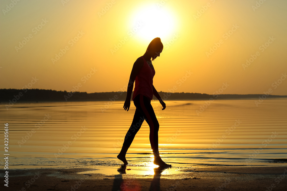 Silhouette of a young beautiful girl walking along the seashore during orange sunset