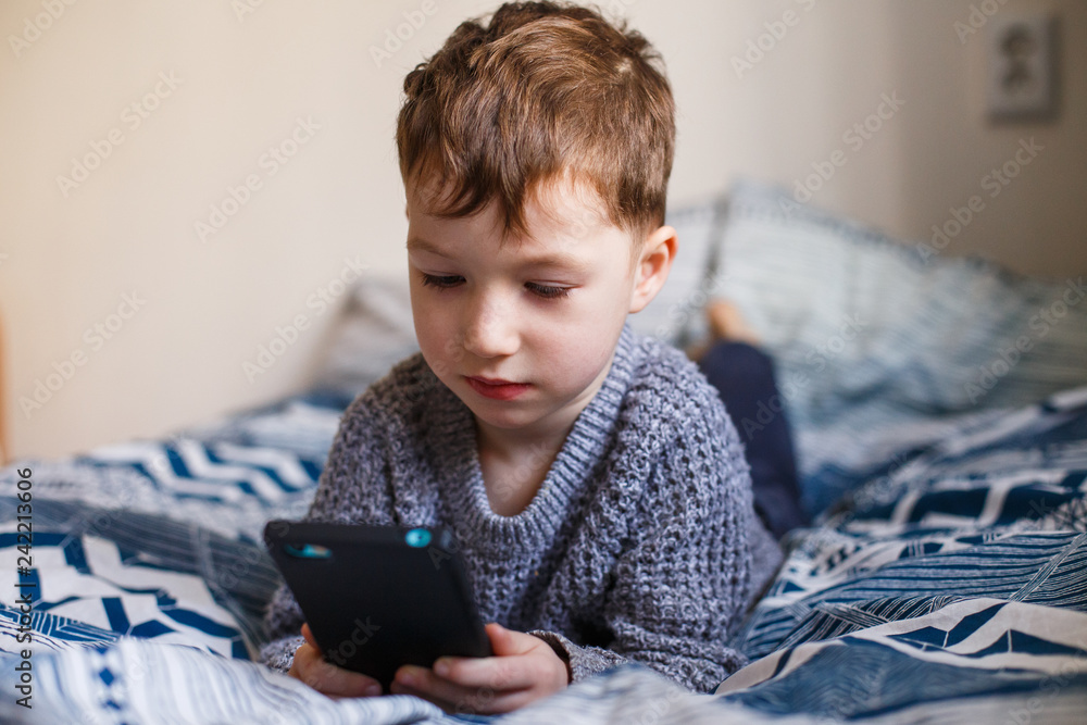 Cute little toddler boy playing with a smartphone on the bed. Internet dependence concept Cute little toddler boy playing with a smartphone on the bed. Internet dependence concept