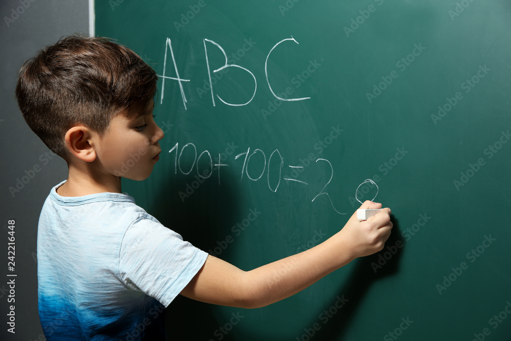 Little child writing letters and doing math on green blackboard