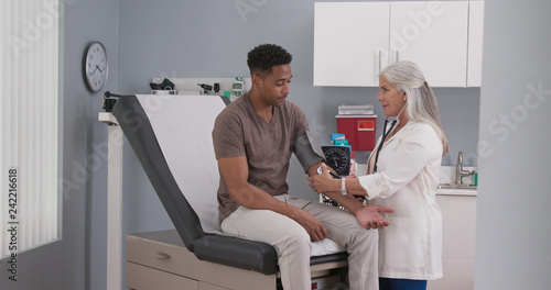 Mature female medical practitioner measuring blood pressure with monitor and stethoscope of a young male patient. Attractive black man having his blood pressure measured by senior white doctor