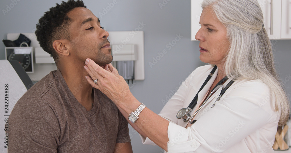 Obraz premium Close up of senior female doctor examining male patients neck. Portrait of attractive black male having doctor checkup at health clinic
