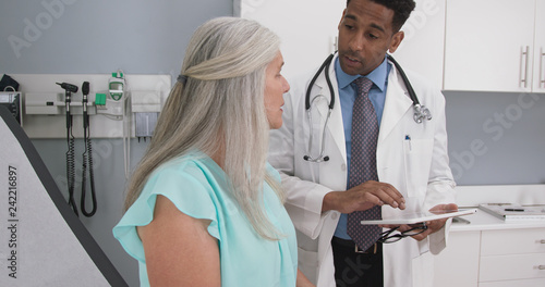 Male millennial MD taking notes on electronic tablet while listening to senior female patient. Elderly woman explaining health condition to young doctor while he uses computer pad