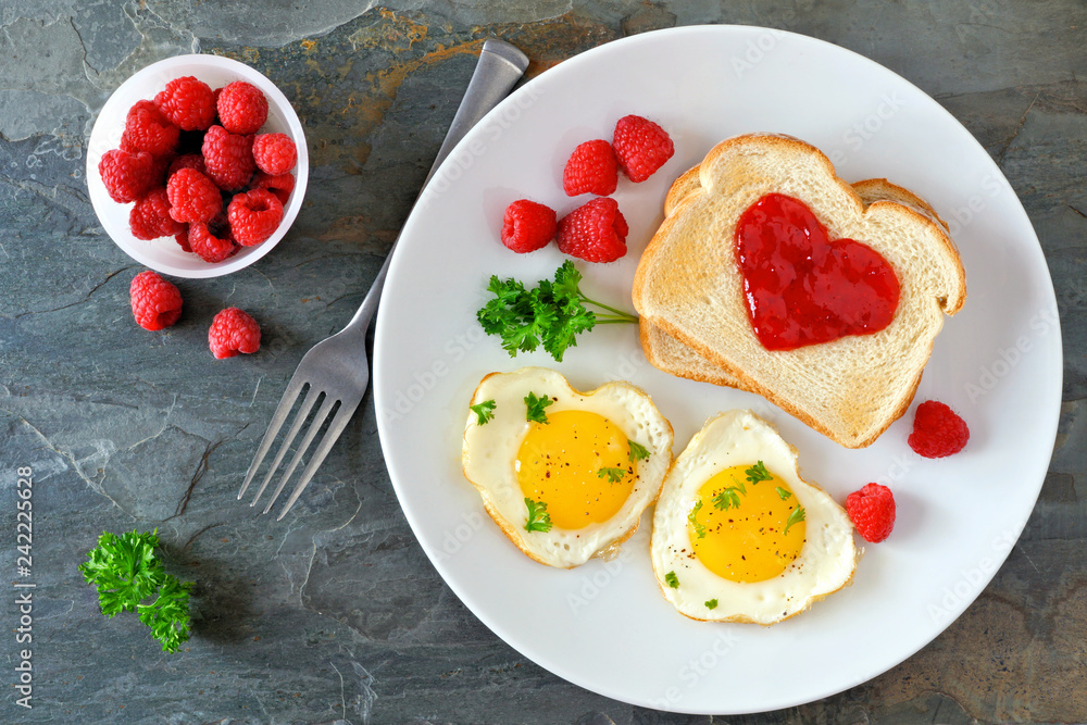 Valentines Day brunch concept with heart shaped eggs and toast with jam over a dark stone background. Top view, table scene. Valentines Day brunch concept with heart shaped eggs and toast with jam over a dark stone background. Top view, table scene.