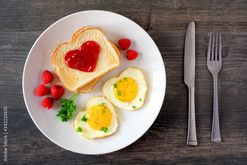 Valentines Day brunch concept with heart shaped eggs and toast with jam over a dark wood background. Top view, table scene. Valentines Day brunch concept with heart shaped eggs and toast with jam over a dark wood background. Top view, table scene.