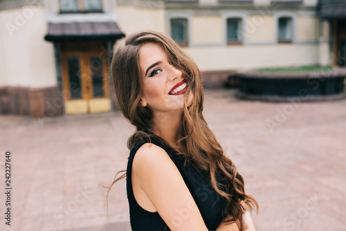 Portrait of pretty girl with long curly hair posing to camera on street on old building background. She wears black dress, vinous lips. Hair covers half her face, she is smiling.