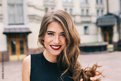 Closeup portrait of effective girl with long curly hair smiling to camera on street on building background. She wears black dress, vinous  lips.