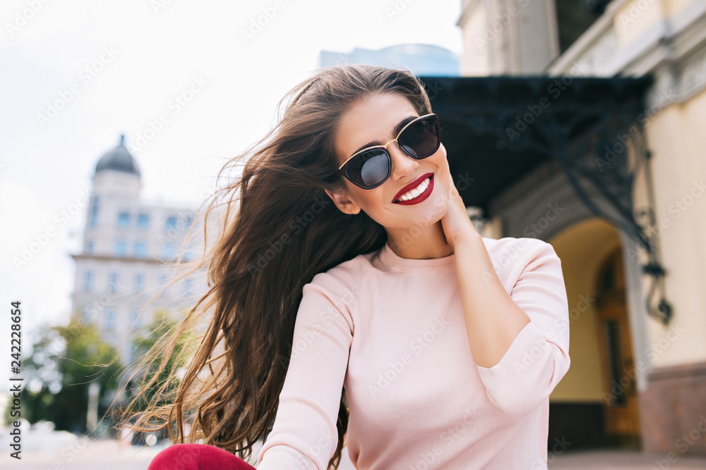 Closeup portrait of attractive girl in sunglasses with vinous lips in the city. Her long hair is flying on wind, she is smiling with snow-white smile to camera.