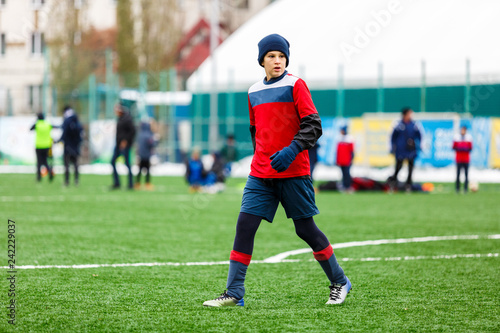 Boy in red and blue sportswear plays soccer on green grass field. Youth football game. Children sport competition, kids plays outdoor, winter activities, training