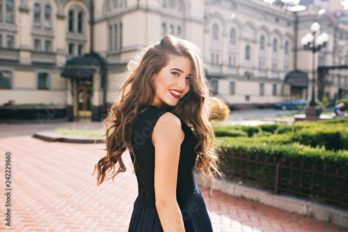 Portrait from back of elefant girl with long curly hair walking on steer on old building background. She has black dress and red lips. She is smiling to camera.