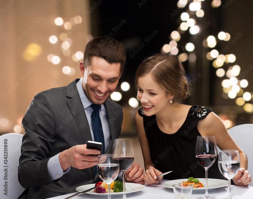 technology, leisure and people concept - happy couple with smartphone eating at restaurant over festive lights on background technology, leisure and people concept - happy couple with smartphone eating at restaurant over festive lights on background