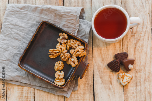 Fototapeta Naklejka Na Ścianę i Meble -  Cup of hot black tea with chocolate candy and walnuts on wooden table. Flat lay, top view.