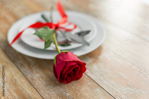 valentines day, table setting and romantic dinner concept - close up of red rose flower on set of dishes with cutlery on wooden background