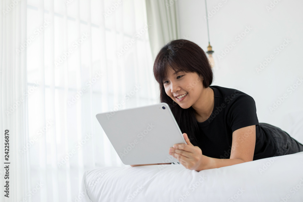 Smiling Asian woman using tablet for video conversation while relaxing on bed in home. Concept of young business people working at home. Copy space. Smiling Asian woman using tablet for video conversation while relaxing on bed in home. Concept of young business people working at home. Copy space.