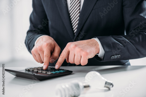 cropped view of businessman in suit using calculator near fluorescent lamps on white background, energy efficiency concept