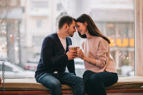 Loving couple dressed in sweaters and jeans is sitting close to each other on the windowsill in a cafe and holding cups in their hands