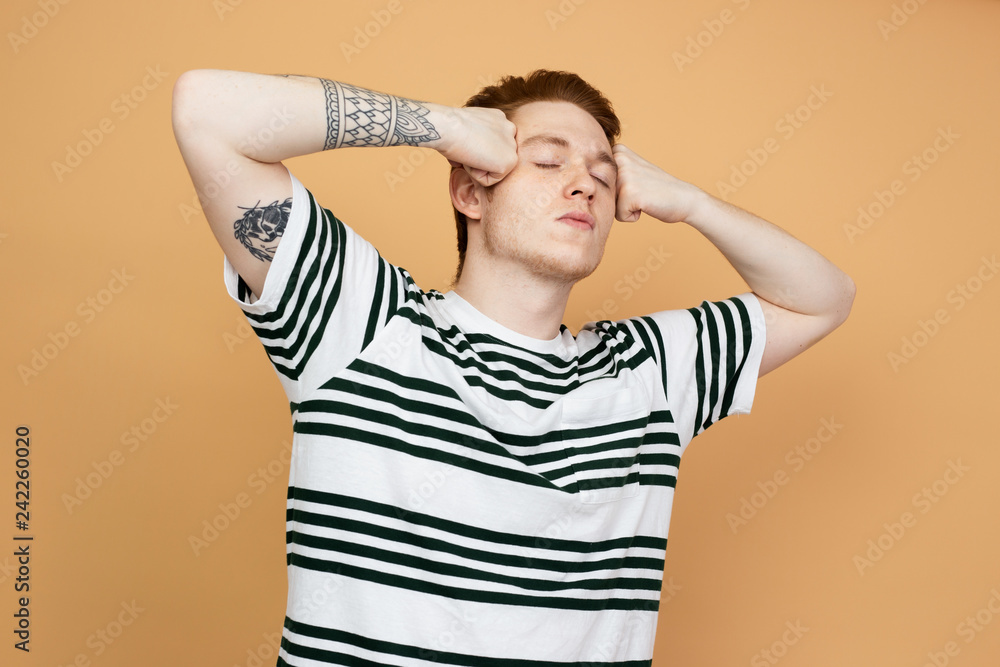 Red-haired stylish guy in a striped shirt with tattoo on his hand is posing holding his hands on his head on the beige background in the studio Red-haired stylish guy in a striped shirt with tattoo on his hand is posing holding his hands on his head on the beige background in the studio