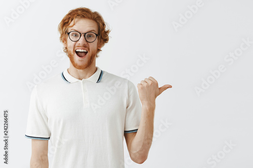 Excited overemotive funny redhead male model with messy beard in polo shirt and glasses pointing right with thumb and smiling joyfully retelling interesting and amazing story happened that way photo