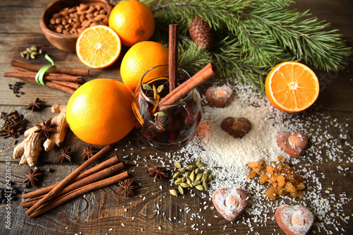 mulled wine in a glass with two sticks of cinnamon and spices on a wooden background with heart-shaped cookies, with a spruce twig, with ingredients for it: cinnamon, anise, cardamom, pepper, oranges
