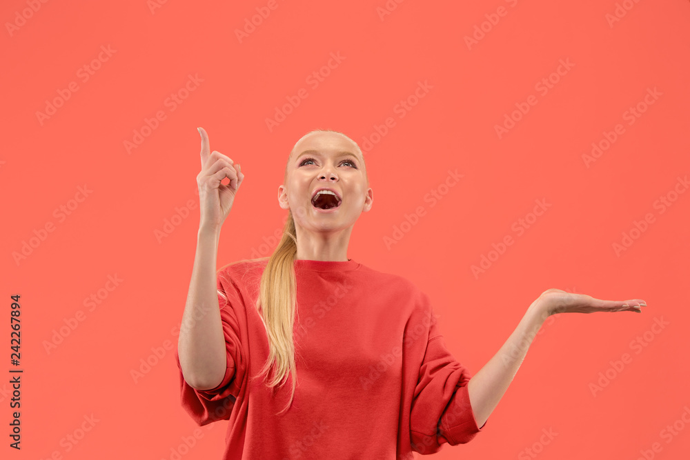Wow. Beautiful female half-length front portrait isolated on coral studio backgroud. Young emotional surprised woman pointing up. Human emotions, facial expression concept. Trendy colors