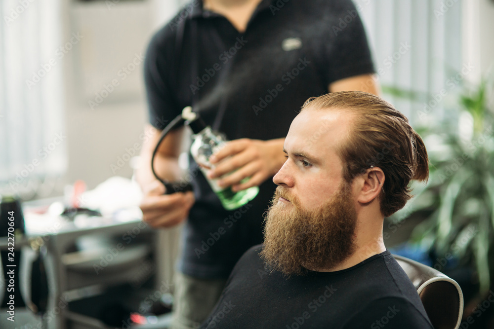Fototapeta premium The hands of young barber making haircut to attractive bearded man in barbershop